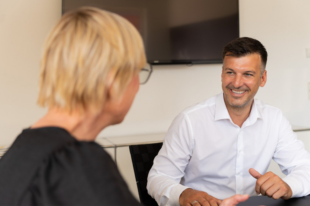 L&auml;chelnder apm-Berater im wei&szlig;en Hemd im Beratungsgespr&auml;ch am Besprechungstisch.