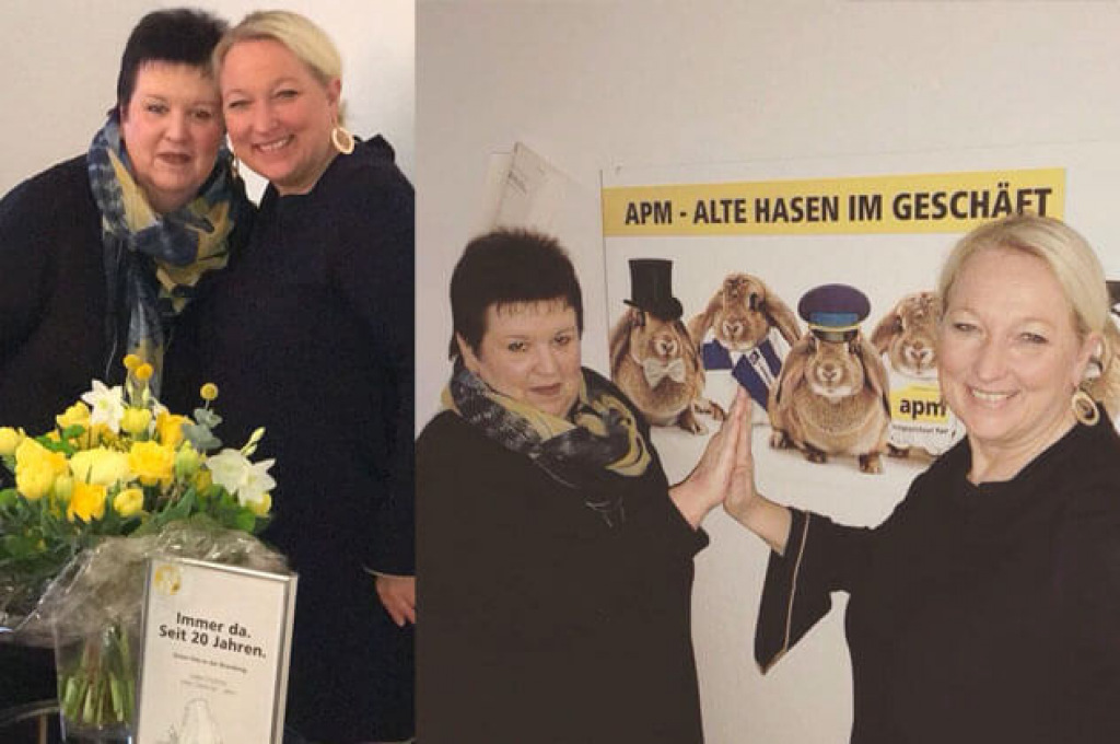 Zwei Kolleginnen mit Blumenstrau&szlig; und Jubil&auml;umsurkunde sowie vor einem Plakat mit dem Motto apm &ndash; alte Hasen im Gesch&auml;ft.