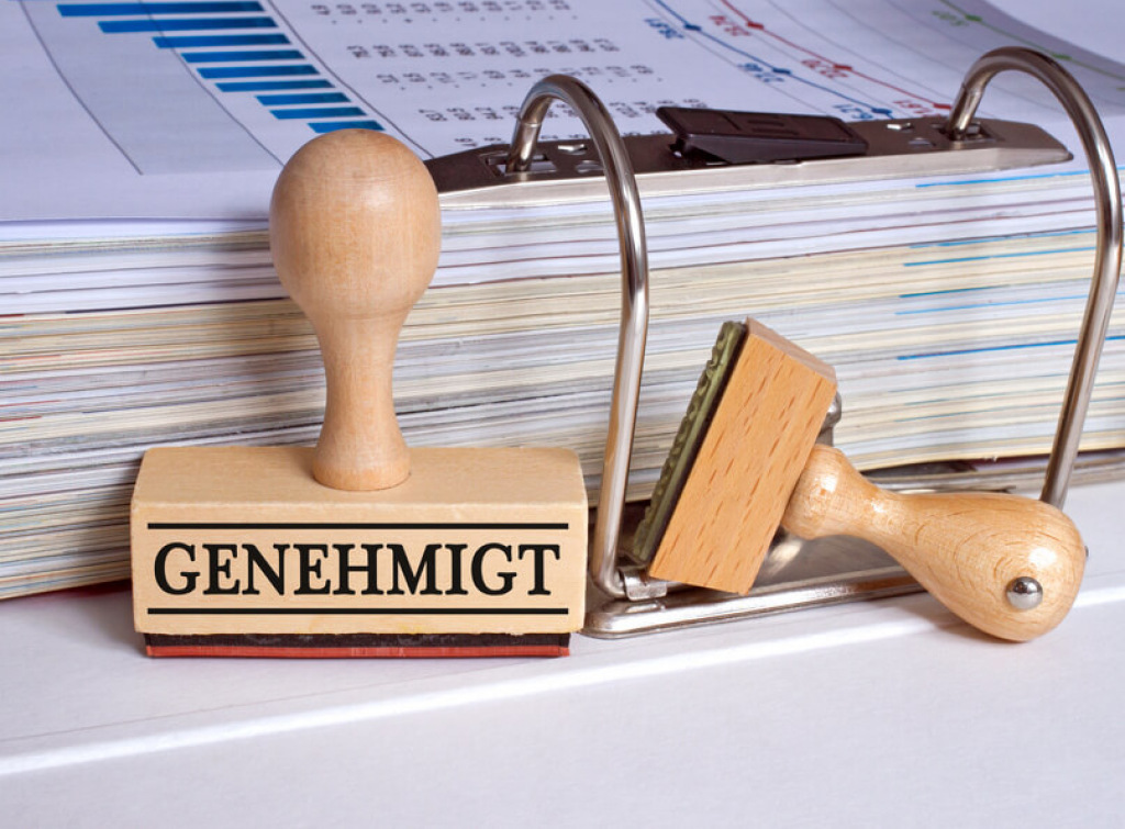 Holzstempel mit der Aufschrift Genehmigt vor einem Aktenordner.