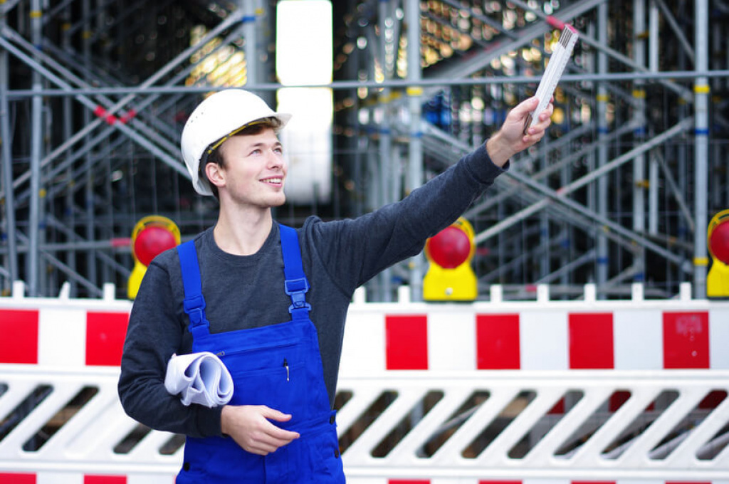 L&auml;chelnder junger Bauarbeiter mit Schutzhelm und Bauplan winkt auf der Baustelle.