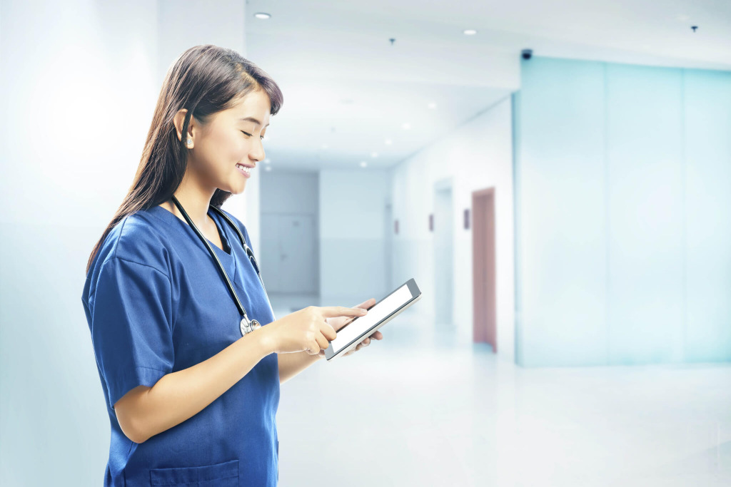 L&auml;chelnde Pflegerin im blauen Kasack mit Stethoskop bedient ein Tablet im Klinikflur.