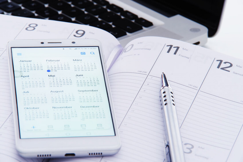 Smartphone mit Jahreskalender auf einem aufgeschlagenen Terminplaner neben Laptop und Stift.